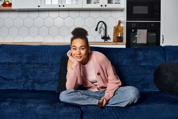 atractiva mujer afroamericana alegre en ropa de casa viendo la televisión mientras está sentado en el sofá en casa - Foto, Imagen