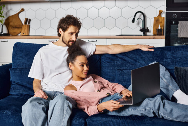 atractiva pareja interracial en acogedora ropa de casa pasar tiempo juntos y ver películas en el portátil - Foto, imagen