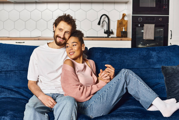 loving joyous multiracial couple in homewear hugging on sofa and smiling happily while at home - Photo, Image