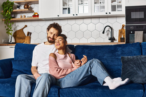 séduisant couple interracial joyeux en tenue décontractée étreignant amoureusement sur le canapé tandis qu'à la maison - Photo, image