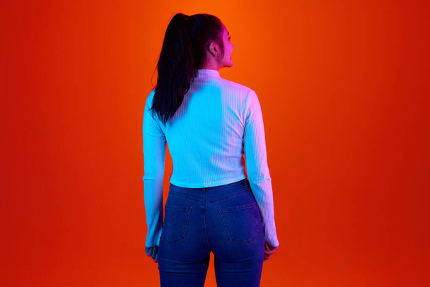 Задний вид изображения женщины в белой рубашке, стоящей, позируя на красно-оранжевом градиентном фоне студии в неоновом свете. Бизнес, уверенность, женская мода. Понятие человеческих эмоций, образа жизни - Фото, изображение