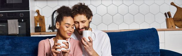 radosna, zróżnicowana para w luźnych strojach pijąca kawę na sofie i ciesząca się sobą nawzajem, sztandar - Zdjęcie, obraz
