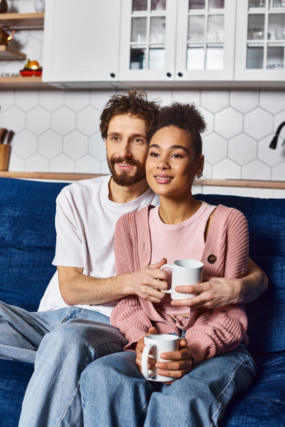 wielorasowy wesoły para w homewear siedzi na kanapie razem i pije gorącą kawę w domu - Zdjęcie, obraz