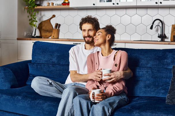 自宅でソファーに座っている間一緒にコーヒーを飲むホームウェアの楽しい異人種間のカップル - 写真・画像