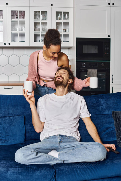 amante multirracial alegre pareja en la ropa de casa beber café y mirándose en casa - Foto, Imagen