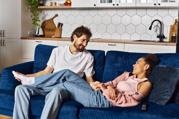handsome jolly man sitting on sofa and looking at his beautiful african american girlfriend - Foto, Imagen