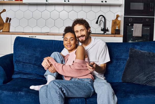 χαρούμενο πολυπολιτισμικό ζευγάρι σε άνετο homewear αγκαλιάζει τον καναπέ και κοιτάζοντας την κάμερα στο σπίτι - Φωτογραφία, εικόνα