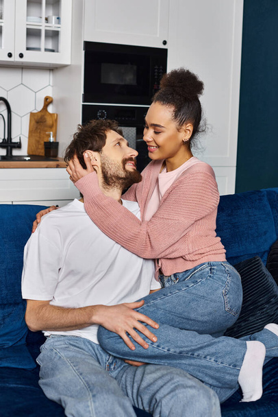 привлекательная межрасовая пара в уютной домашней одежде сидит на диване и счастливо улыбается друг другу - Фото, изображение