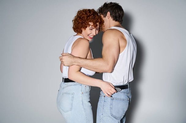 alegre seductor hombre y mujer en jeans posando sobre gris telón de fondo y sonriendo felizmente, sexy pareja - Foto, Imagen