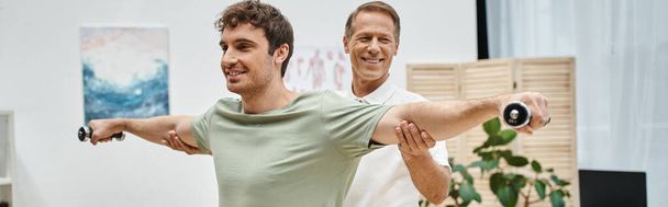 jolly mature doctor helping his cheerful patient to use dumbbells during rehabilitation, banner - Photo, Image