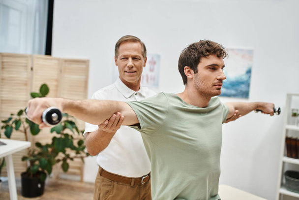 cheerful mature doctor in uniform helping his handsome patient to use dumbbells on rehabilitation - Photo, Image