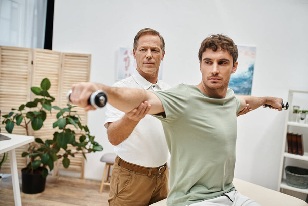 attractive mature doctor helping his handsome patient to use dumbbells on rehabilitation in ward - Photo, Image