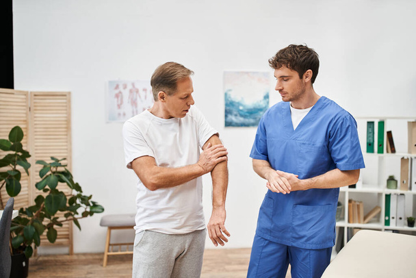 красивый преданный врач в синей форме слушает своего взрослого пациента во время приема - Фото, изображение