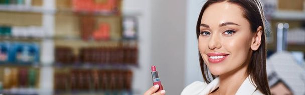 joyous good looking woman in business casual choosing lipstick and smiling at camera, banner - Photo, Image