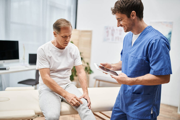 весёлый красивый доктор в синем халате держит планшет и слушает своего взрослого пациента в больнице - Фото, изображение