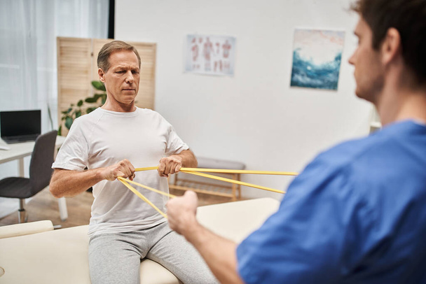 focus on mature patient training with resistance band with his blurred doctor, rehabilitation - Photo, Image