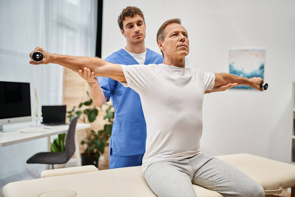 good looking mature patient in casual attire using dumbbells with help of his handsome doctor - Photo, Image