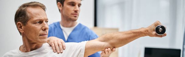 toegewijde arts in blauwe badjas helpen volwassen aantrekkelijke patiënt met halter tijdens revalidatie - Foto, afbeelding