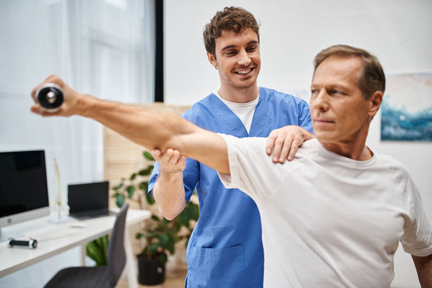 médico alegre en bata azul ayudando a su paciente guapo maduro a usar mancuerna durante la rehabilitación - Foto, Imagen
