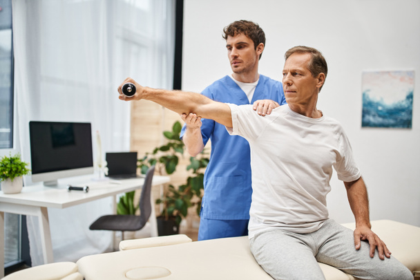 good looking doctor in blue robe helping his mature handsome patient with dumbbell on rehabilitation - Photo, Image