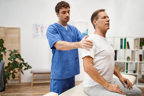 εμφανίσιμος γιατρός με μπλε στολή βοηθά τον ώριμο ασθενή του να αποκαταστήσει τους μυς του στην πτέρυγα. - Φωτογραφία, εικόνα