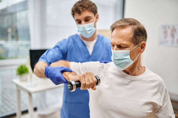 odaadó jóképű orvos maszkkal és kesztyűvel, aki segít a betegének súlyzókat használni a találkozón. - Fotó, kép