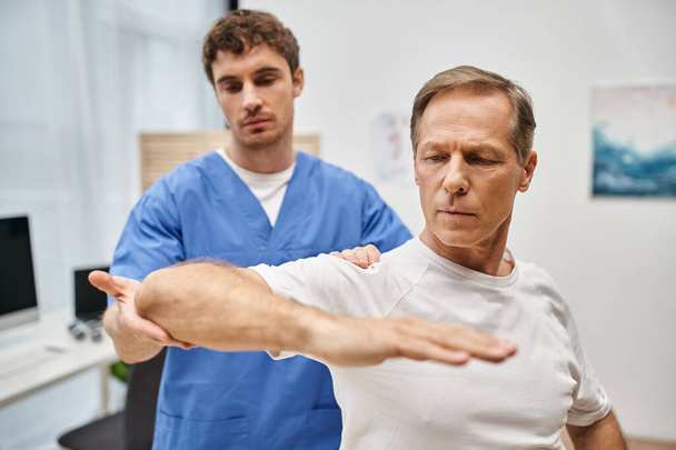 εμφανίσιμος αφοσιωμένος γιατρός με μπλε ρόμπα βοηθά τον ασθενή του να αποκαταστήσει το σώμα του στην πτέρυγα. - Φωτογραφία, εικόνα