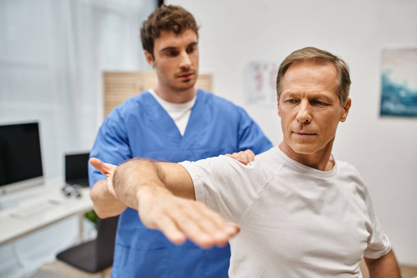 atraktivní tvrdě pracující lékař v modrém rouchu pomáhá svému pacientovi rehabilitovat jeho tělo v oddělení - Fotografie, Obrázek