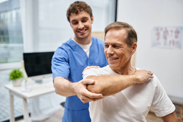 веселий привабливий лікар, який допомагає своєму зрілому пацієнту розтягуватися під час реабілітації в лікарні - Фото, зображення