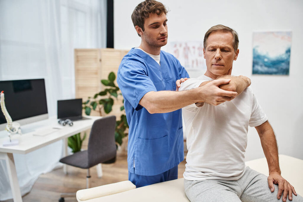 mature handsome patient in casual attire stretching his muscles with help of his attractive doctor - Photo, Image