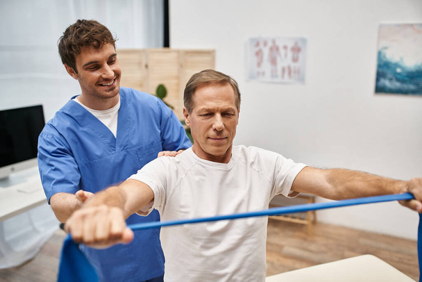 joyous attractive doctor in blue robe helping his mature patient to use resistance band in hospital - Photo, Image