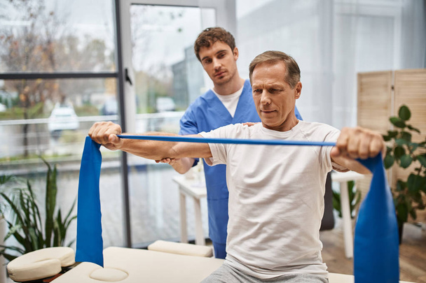 трудолюбивый врач в синей форме помогает своему взрослому пациенту с полосой сопротивления на реабилитации - Фото, изображение