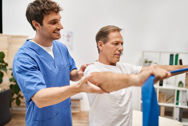 joyeux beau médecin en robe bleue aidant son patient mature à utiliser la bande de résistance à l'hôpital - Photo, image