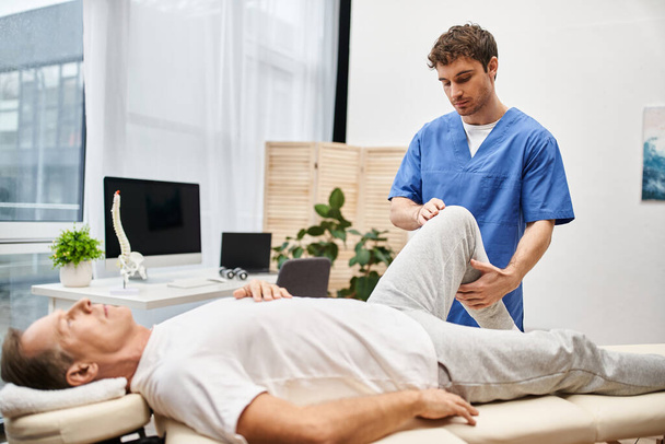 good looking doctor in blue robe helping his mature patient to stretch during rehabilitation - Photo, Image