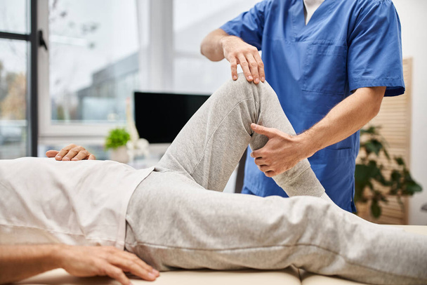 обрезанный вид преданного врача в синем халате растягивающего мышцы своего взрослого пациента в больнице - Фото, изображение