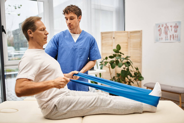 good looking dedicated doctor helping his mature patient to use resistance band, rehabilitation - Photo, Image