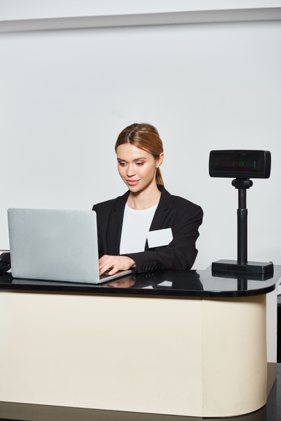 ελκυστική κομψή πωλήτρια με μαύρο σακάκι που εργάζεται σε φορητό υπολογιστή, ενώ κάθεται σε πάγκο στο κατάστημα - Φωτογραφία, εικόνα