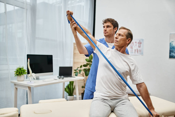 handsome dedicated doctor helping mature patient to use resistance band on rehabilitation in ward - Photo, Image