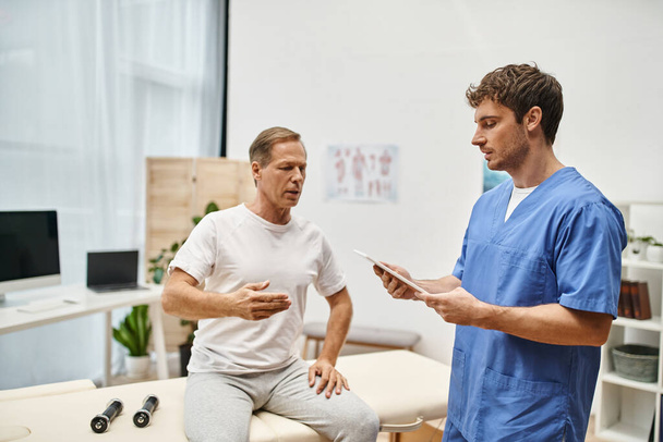 goed uitziende arts in blauw gewaad die tablet vasthoudt tijdens een afspraak met zijn volwassen knappe patiënt - Foto, afbeelding