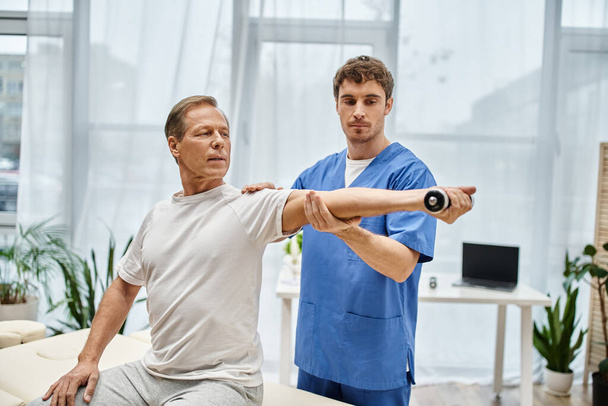 красивий лікар в блакитному халаті допомагає своєму зрілому пацієнту в повсякденному вбранні використовувати гантелі в лікарні - Фото, зображення