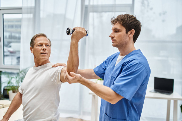 hardworking doctor helping his mature patient in casual attire to use dumbbell on appointment - Photo, Image