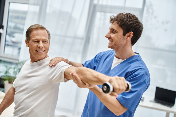 knappe vrolijke dokter helpen zijn vrolijke volwassen patiënt te gebruiken halter tijdens de revalidatie - Foto, afbeelding