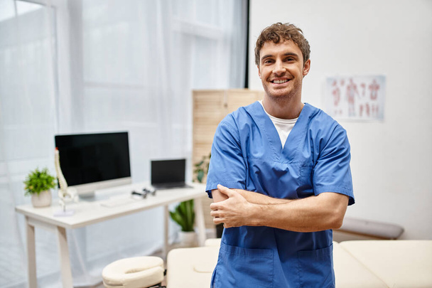 iloinen omistautunut kuntoutuslääkäri sinisessä univormussa poseeraa sairaalaosastolla ja hymyilee kameralle - Valokuva, kuva
