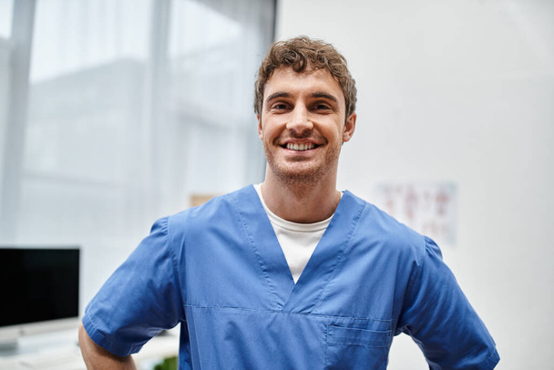 vrolijke goed uitziende rehabilitoloog in blauw uniform poseren in ziekenhuis afdeling en glimlachen op camera - Foto, afbeelding
