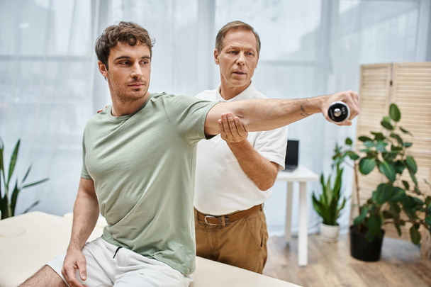 mature hardworking doctor helping his appealing patient to use dumbbell during rehabilitation - Photo, Image
