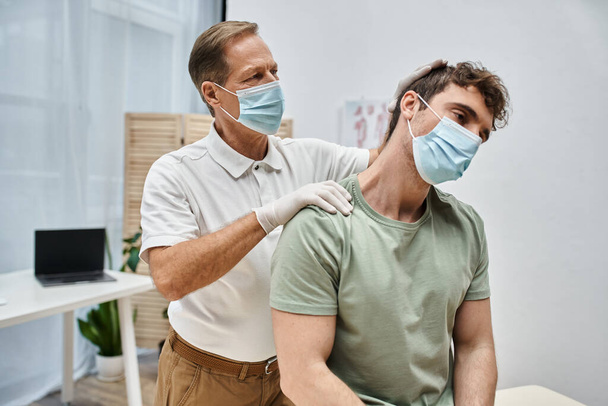omistautunut kuntoutuslääkäri, jolla on maski ja käsineet potilaan hierova kaula sairaalaosastolla - Valokuva, kuva
