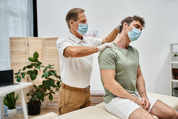 dedicado bonito maduro médico no uniforme com máscara massageando pescoço de seu paciente no hospital - Foto, Imagem