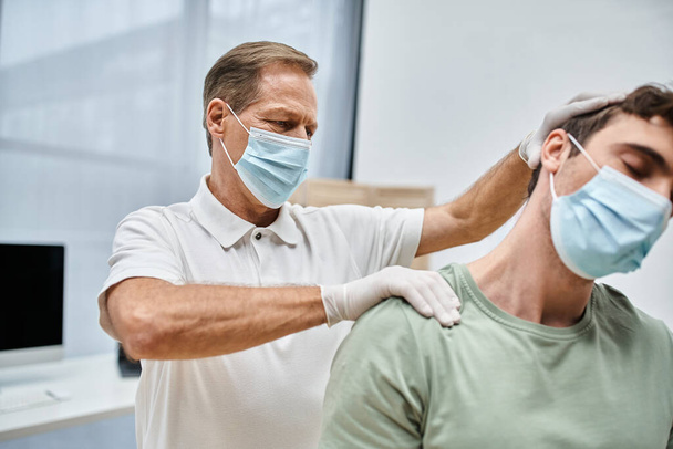 hardwerkende volwassen arts met masker en handschoenen masseren nek van zijn patiënt in ziekenhuis afdeling - Foto, afbeelding