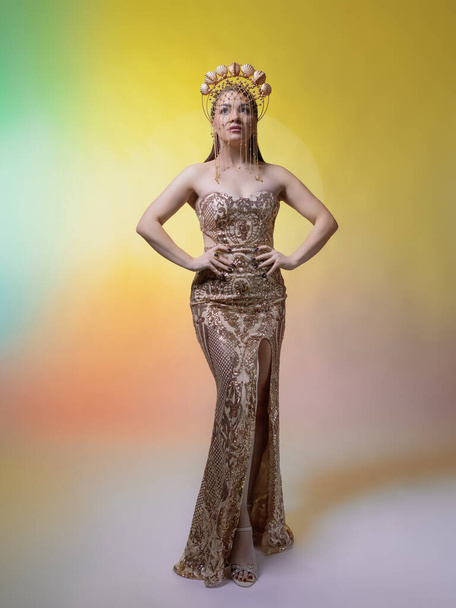 Piękna, szczupła kobieta w koronie muszelek pokrytej złotem. Ma na sobie obcisłą sukienkę z błyszczących pływaków. Na żółtym tle - Zdjęcie, obraz
