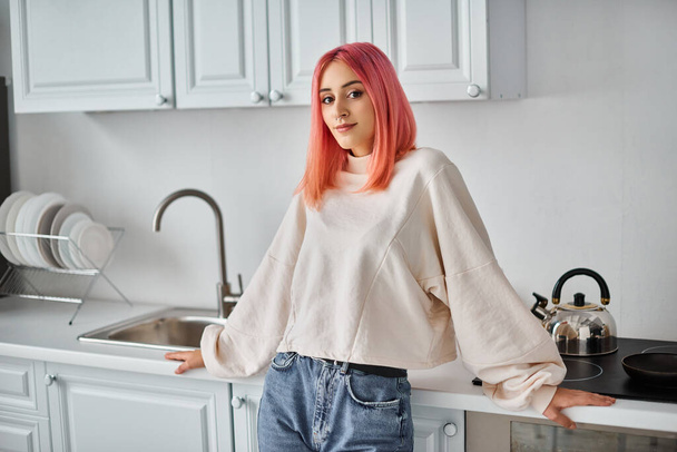 cheerful attractive woman in white casual jumper and jeans looking at camera while in kitchen - Photo, Image